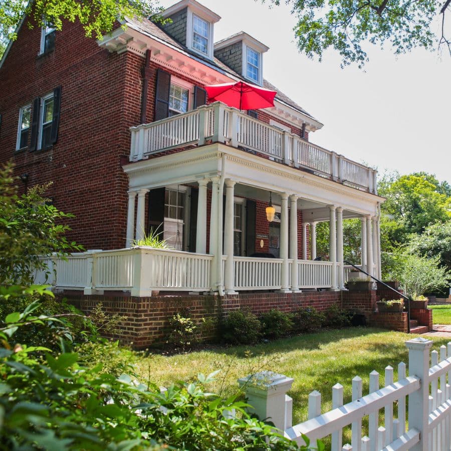 A brick house with a porch and a red umbrella overlooking a green lawn and white picket fence.