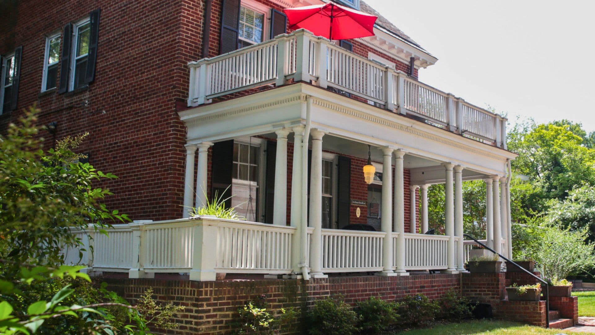 A brick house with a white porch and a red umbrella overhead.