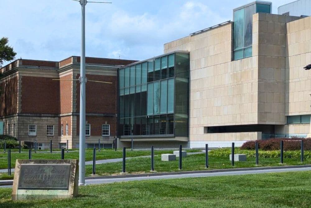 A modern building with glass elements next to a brick structure, surrounded by green grass and a memorial stone.