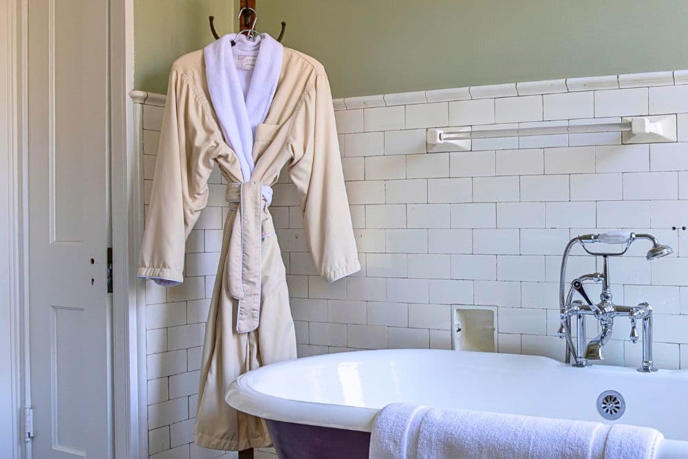 A cream-colored robe hangs beside a white bathtub in a tiled bathroom.