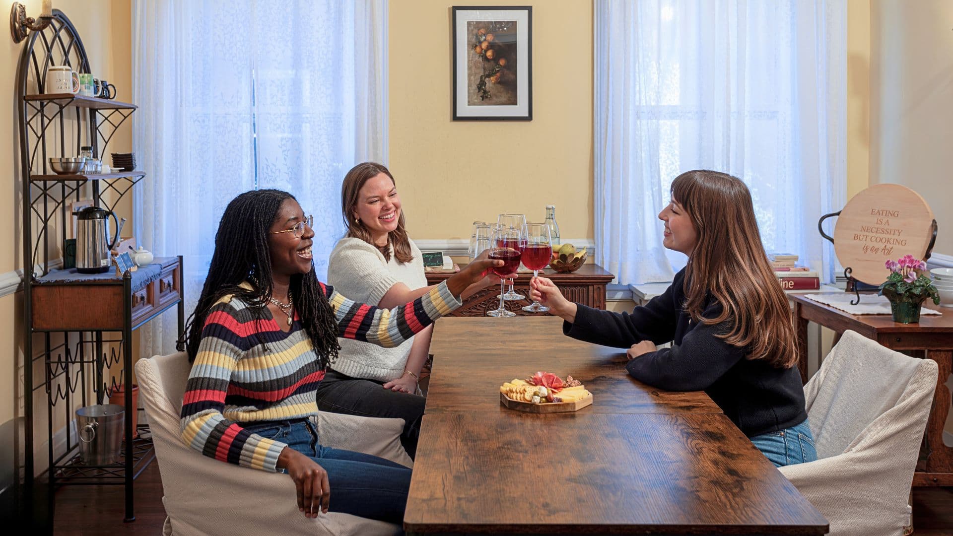 Three women toast with glasses of red wine at a wooden dining table adorned with a charcuterie board.