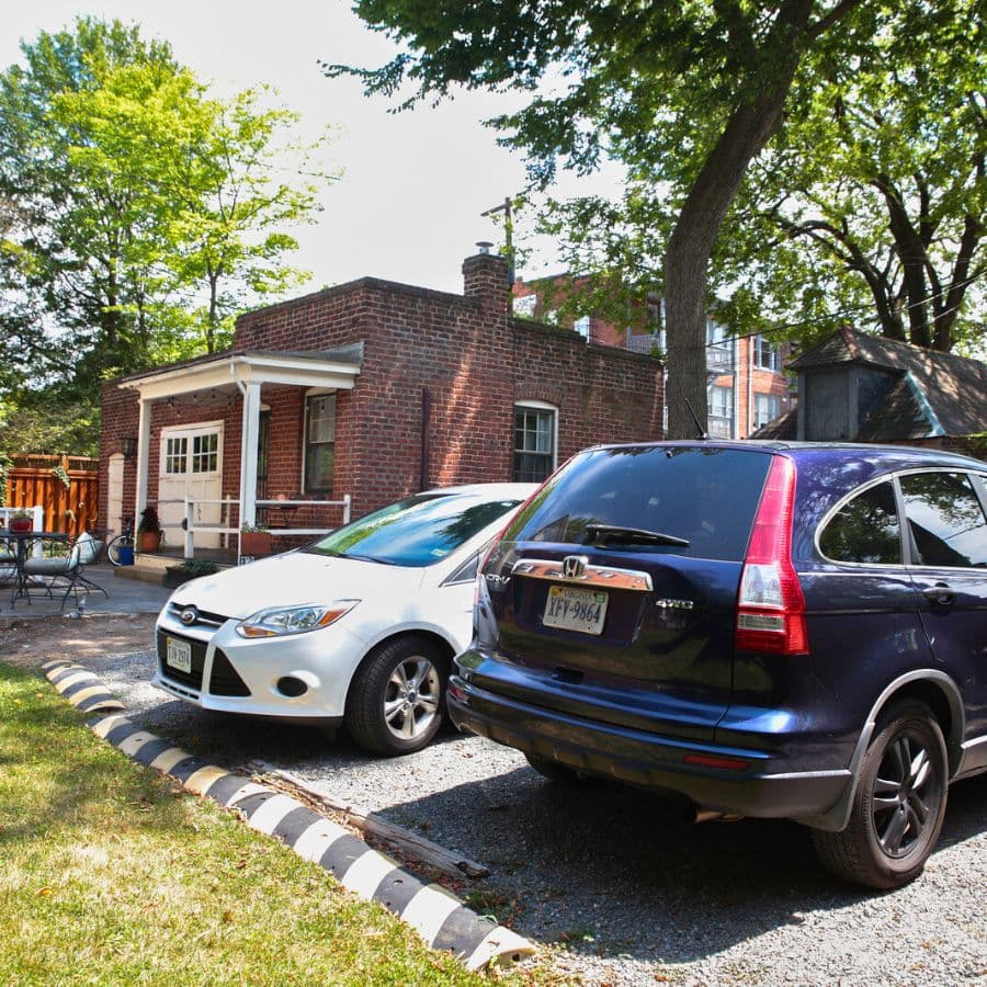 Two parked cars, a white sedan and a dark SUV, beside a brick building and trees.